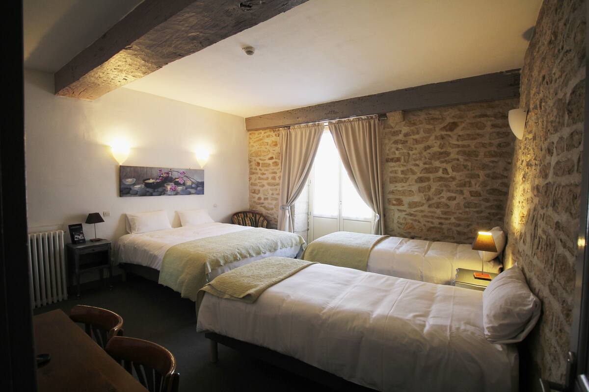 Quarto com duas camas, uma mesa de madeira e uma cadeira. Há um quadro na parede, um abajur na mesa de cabeceira e uma janela com cortinas.
