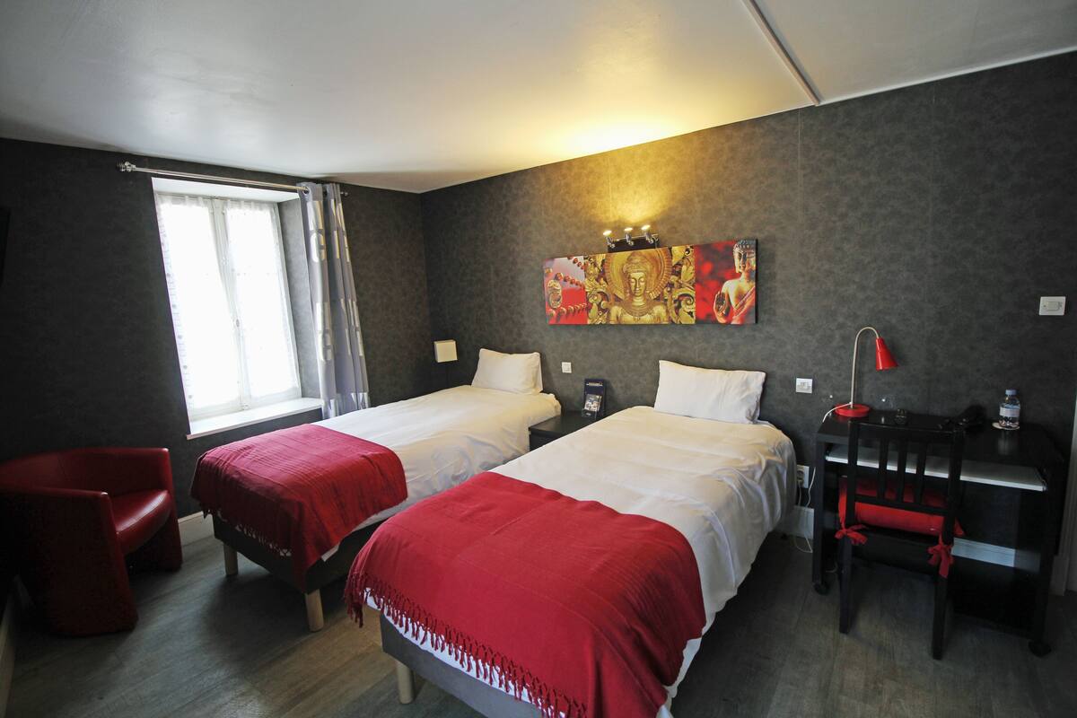 Quarto de hotel com duas camas, uma cadeira vermelha, uma mesa com luminária e um quadro na parede.