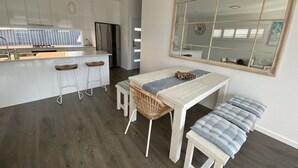 A modern kitchen with white cabinetry, a central island with stools, a dining table with a bench, and a large mirror on the wall. - Modern Beach Getaway  (Shellharbour)