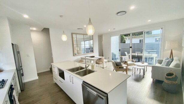 A modern kitchen with a central island, built-in appliances, and a dining area with a glass door to the outside. - Modern Beach Getaway  (Shellharbour)