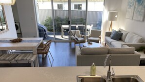 A modern kitchen with a stainless steel sink, a dining area with a table and chairs, a sofa, and a view through large sliding glass doors to an outdoor patio. - Modern Beach Getaway  (Shellharbour)