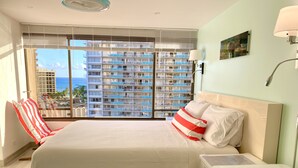 A hotel room with a large bed, a chair, a nightstand, and a view of a high-rise building with balconies. - Discovery Bay-Resolution Ocean View Condo (Honolulu)