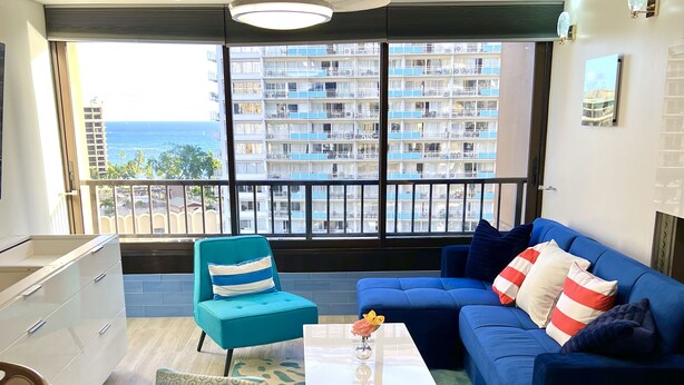 A modern living room with a blue sofa, teal armchairs, and a coffee table. A balcony with a view of a high-rise building and the ocean is visible through large windows. - Discovery Bay-Resolution Ocean View Condo (Honolulu)