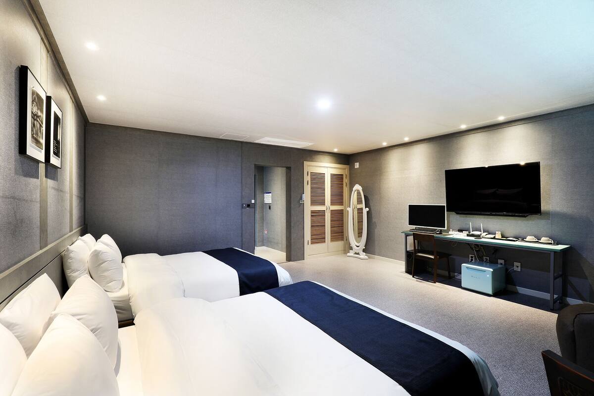 A modern hotel room with two beds, a desk with a computer, a flat-screen TV, and a minibar.