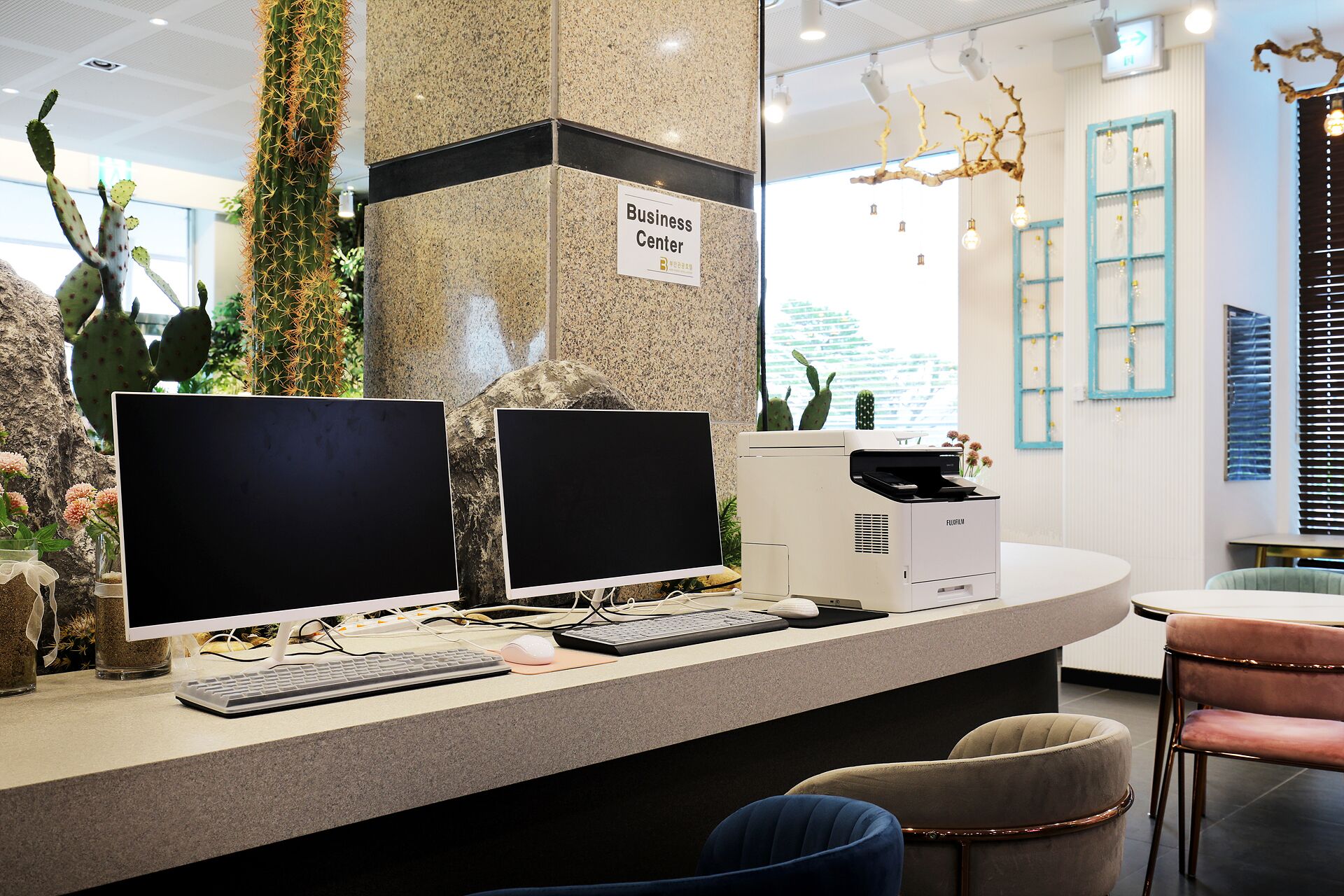 Un comptoir d’accueil équipé de deux écrans, d’un clavier et d’une souris. Une pancarte indique « Centre d’affaires ». Le comptoir est fait d’un matériau à motifs et on y trouve des chaises et une table à proximité.