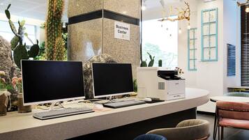 Un comptoir d’accueil équipé de deux écrans, d’un clavier et d’une souris. Une pancarte indique « Centre d’affaires ». Le comptoir est fait d’un matériau à motifs et on y trouve des chaises et une table à proximité.