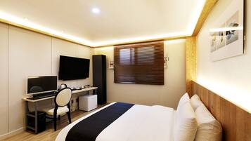 A hotel room with a bed, a desk with a computer, a TV, and a window with blinds.