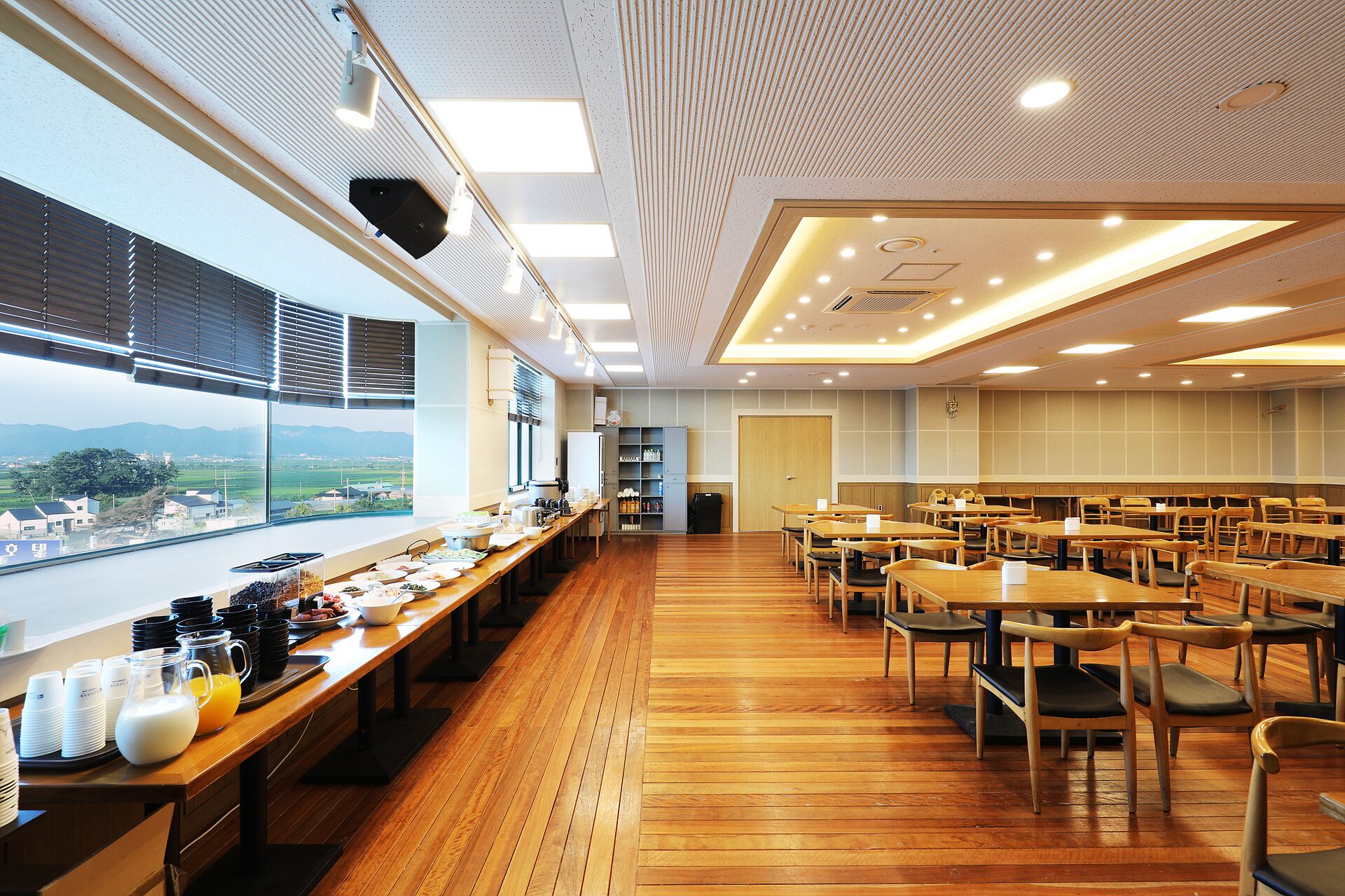 A spacious dining area with long tables and chairs, a buffet setup on the left, and a view of the outdoors through large windows.