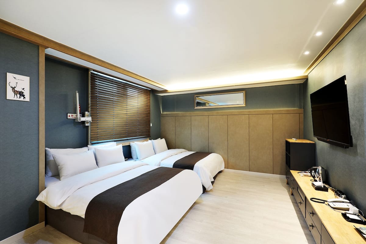A hotel room with a bed, bedside table, TV, and a window with blinds.