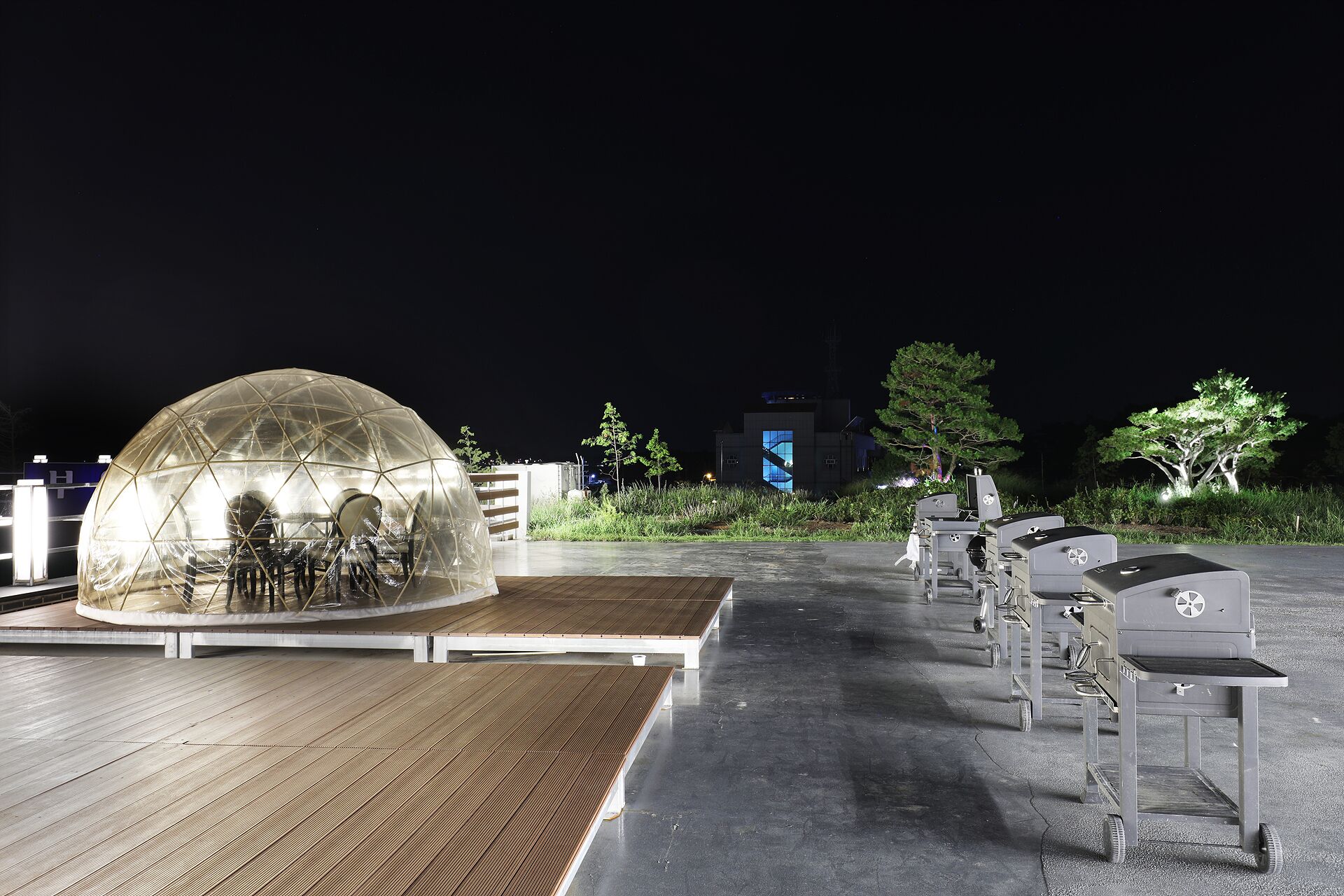 A transparent dome structure with seating inside, a wooden deck, and outdoor grills in a nighttime setting.