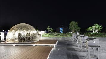 A transparent dome structure with seating inside, a wooden deck, and outdoor grills in a nighttime setting.