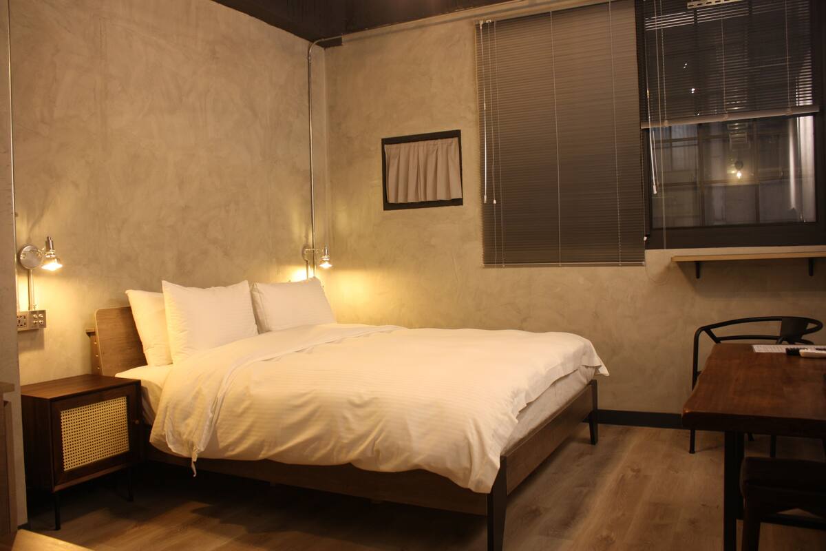 Quarto com cama, luminárias de cabeceira, uma mesinha e janela com persianas.