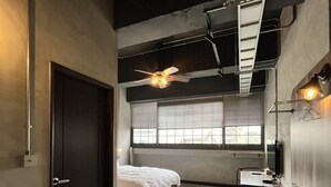 Une chambre d’hôtel de taille réduite, comprenant un lit, un bureau, une chaise, une petite table, un ventilateur de plafond et une fenêtre avec des stores.