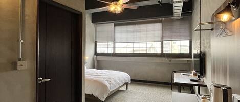 A compact hotel room with a bed, a desk, a chair, a small table, a ceiling fan, and a window with blinds.