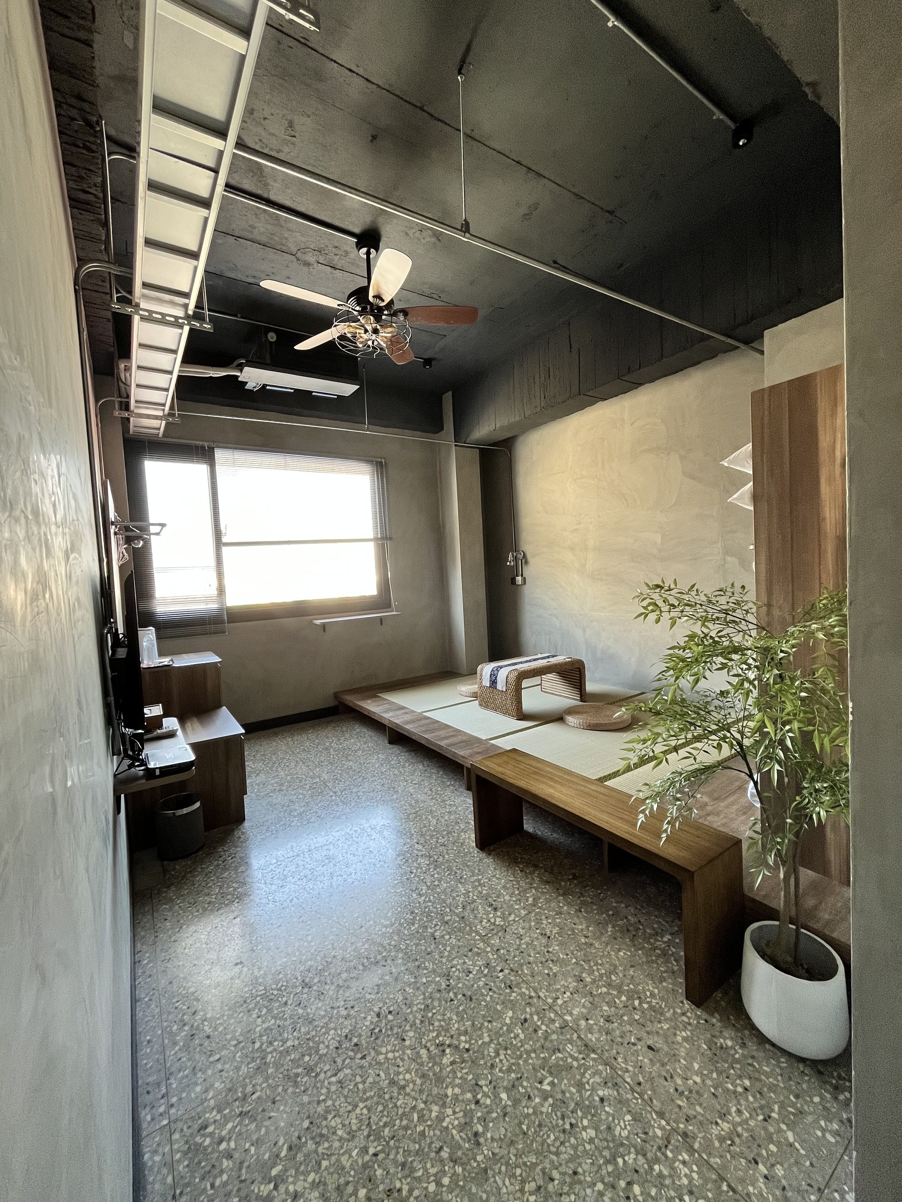 A modern, minimalist room with a concrete ceiling, a ceiling fan, a large window, a wooden bench, a small table, and a potted plant.