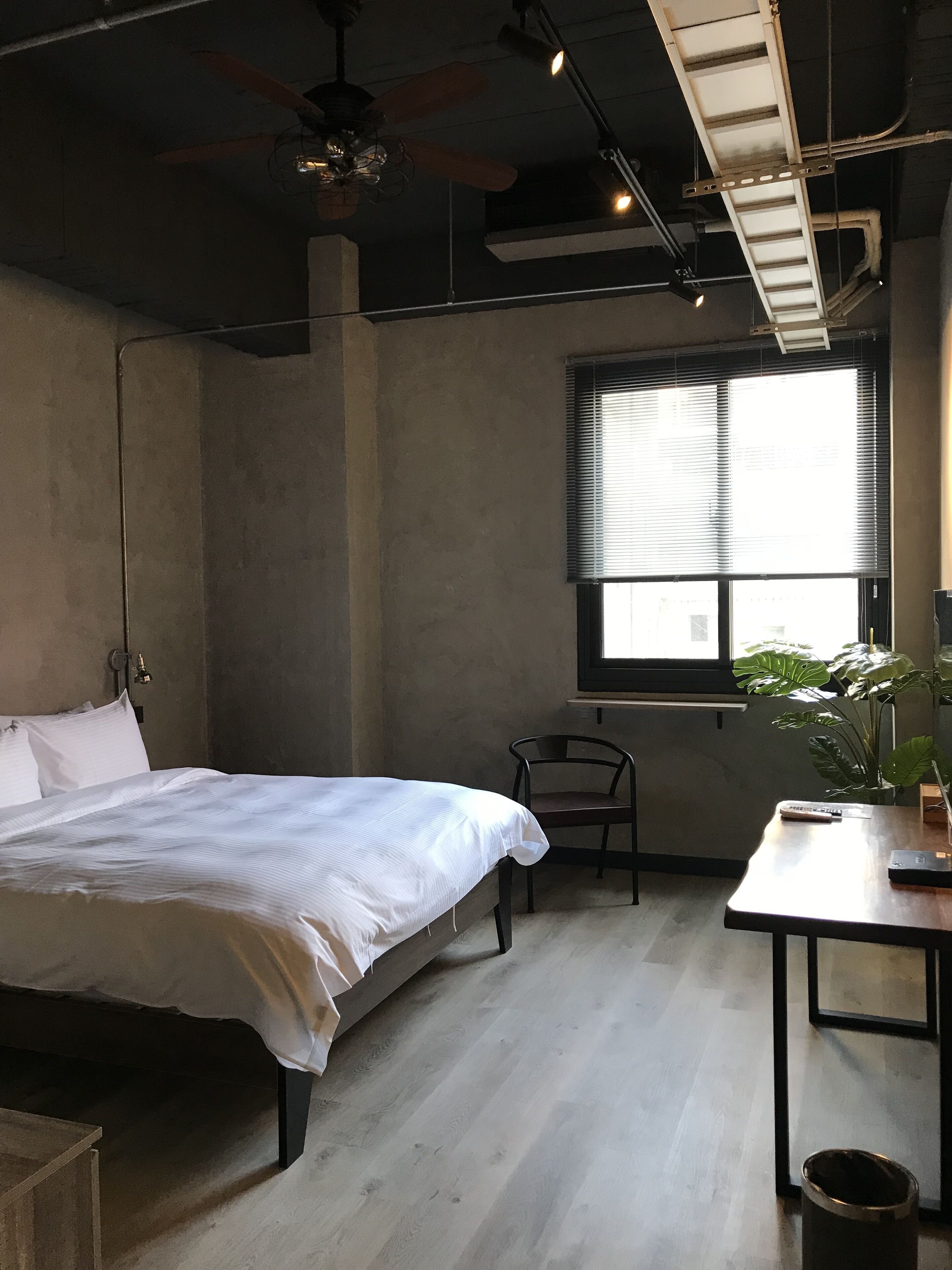 Une chambre à coucher comprenant un lit, une chaise, une table, une fenêtre avec des stores et un ventilateur de plafond.