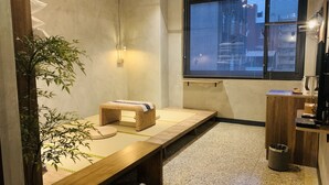 Une petite pièce accueillante, avec un sol recouvert de tatamis, un banc bas en bois, un petit tabouret en osier, une plante de bambou en pot, une fenêtre avec des stores et une armoire en bois contenant une poubelle.