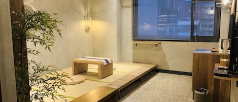 A small, cozy room with a tatami mat floor, a low wooden bench, a small woven stool, a potted bamboo plant, a window with blinds, and a wooden cabinet with a trash can.