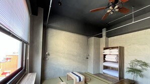 Une chambre moderne dotée d’une grande fenêtre, d’un ventilateur de plafond, d’une étagère intégrée où sont rangées les serviettes, d’un petit banc et d’un sol recouvert de tatamis.