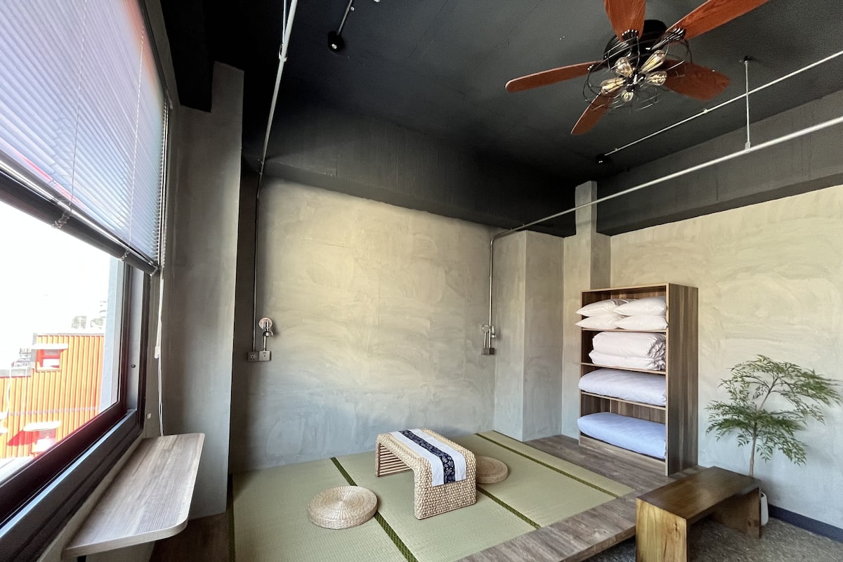 Quarto moderno com uma janela grande, ventoinha no teto, prateleira embutida com toalhas, um pequeno banco e piso de esteira de tatame.