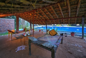 An outdoor seating area with a thatched roof, a pool in the background, and a brick wall. - Bagh Serai (Sawai Madhopur)