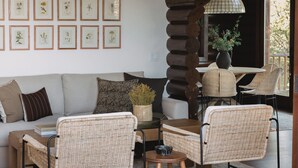 En hyggelig opholdsstue med sofa, fletstole, sofabord og spisebord. Væggen er dekoreret med indrammede plantetryk, og der står en potteplante på bordet.