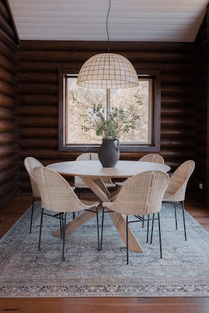 A wooden cabin interior with a round dining table, wicker chairs, a large window, and a hanging light fixture.