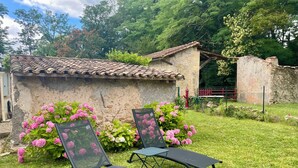 A rustic stone building with a tiled roof, surrounded by lush greenery and pink flowers. Two black metal chairs are placed in front of the building.