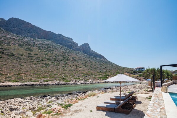 Ein Poolbereich mit Liegestühlen und Sonnenschirmen, eine felsige Küstenlinie und ein Berg im Hintergrund.