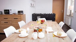 Een eetruimte met een houten tafel gedekt voor het ontbijt, witte stoelen, een zwarte bank, een witte radiator en een houten kast met keukenapparatuur.
