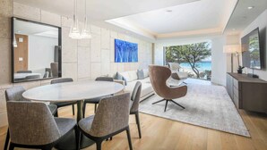 A modern living room with a round dining table, grey upholstered chairs, a large mirror, a blue abstract painting, a sofa with cushions, a unique brown chair, a floor lamp, and a view of the ocean through large windows.