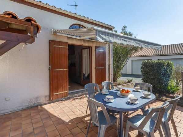 Eine Terrasse mit rundem Tisch und Stühlen, einer Holztür und einem überdachten Bereich mit weißem Sonnensegel.