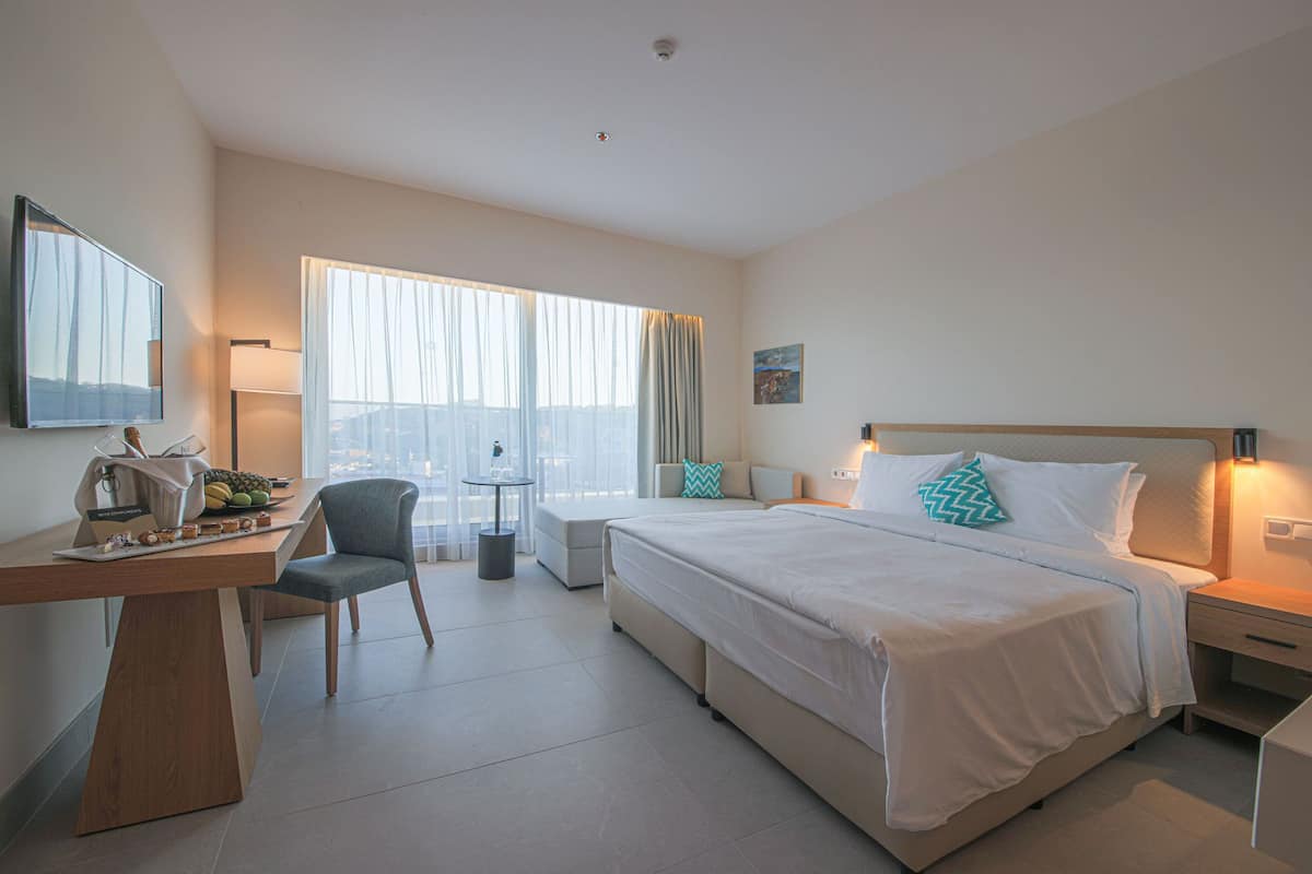A hotel room with a bed, desk, chair, and a view of the city through large windows.