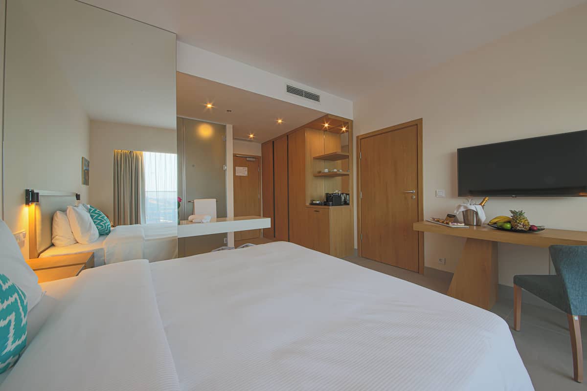 A hotel room with a large bed, a desk with a flat-screen TV, a chair, a wardrobe, and a small kitchenette.