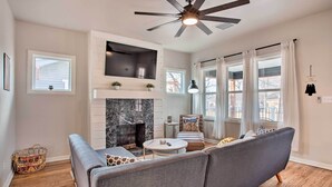 A modern living room with a fireplace, a ceiling fan, and a large window with curtains. - Charming Downtown Home w/ Updated Interior! (Oklahoma City)