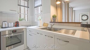 A modern kitchen with white cabinetry, stainless steel appliances, and a large window with blinds. - Super calm cosy & cute 2 bdr at the heart of Old Honfleur- Duplex Le Pélican ★★★★ (Honfleur)