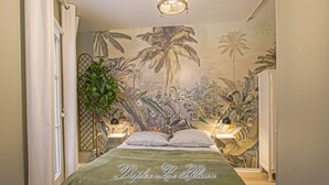 A bedroom with a bed, a mural of tropical plants, a hanging light fixture, and a plant beside the bed. - Super calm cosy & cute 2 bdr at the heart of Old Honfleur- Duplex Le Pélican ★★★★ (Honfleur)