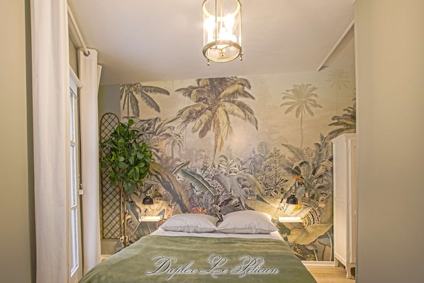 A bedroom with a bed, a mural of tropical plants, a hanging light fixture, and a plant beside the bed.