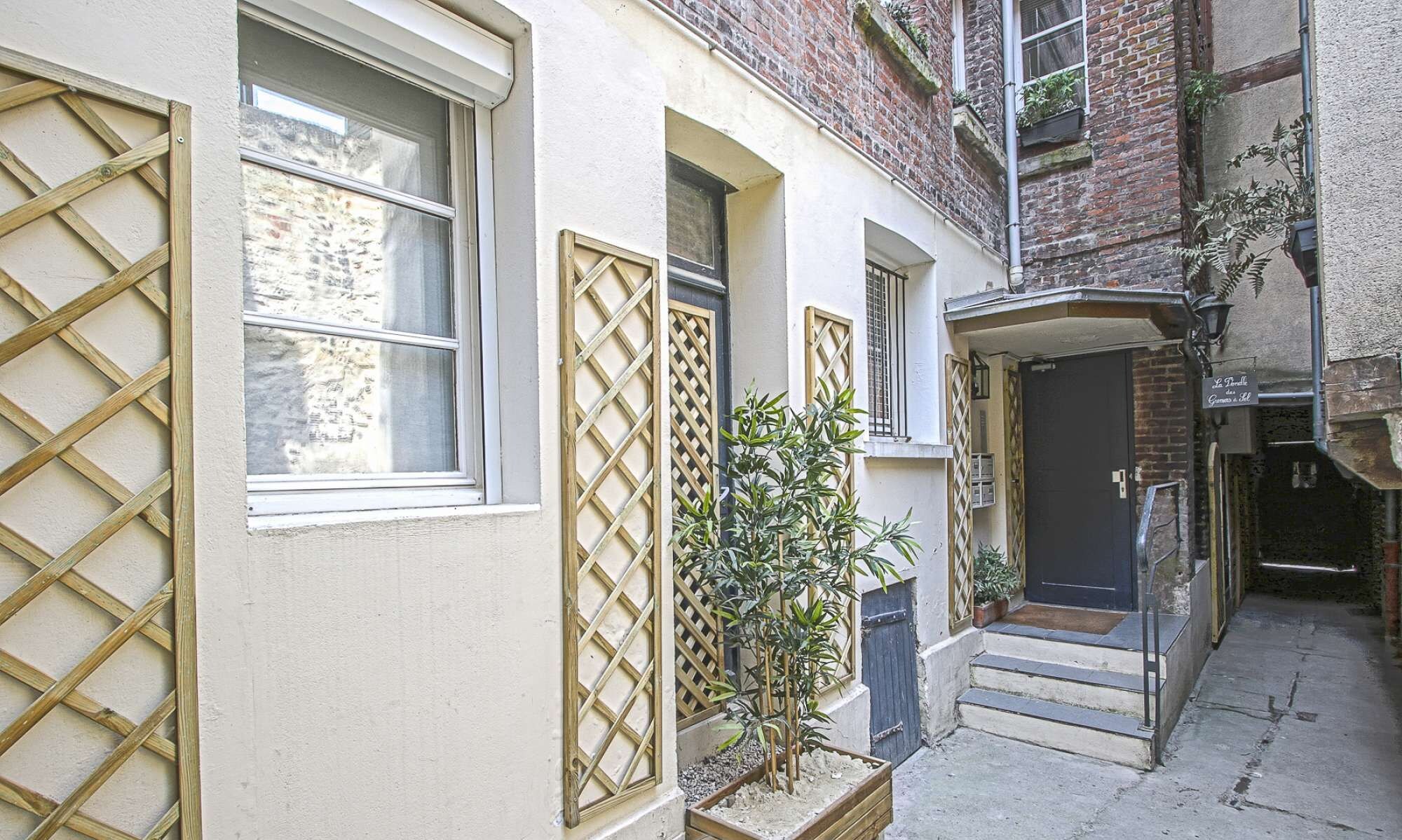 A narrow alleyway with a building featuring a wooden lattice window, a small potted plant, and a door with a sign.