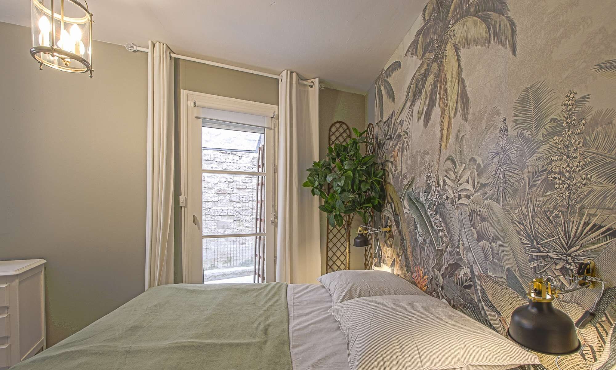 A bedroom with a bed, a window with curtains, a wall mural, a plant, and a lamp.