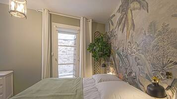 A bedroom with a bed, a window with curtains, a wall mural, a plant, and a lamp.