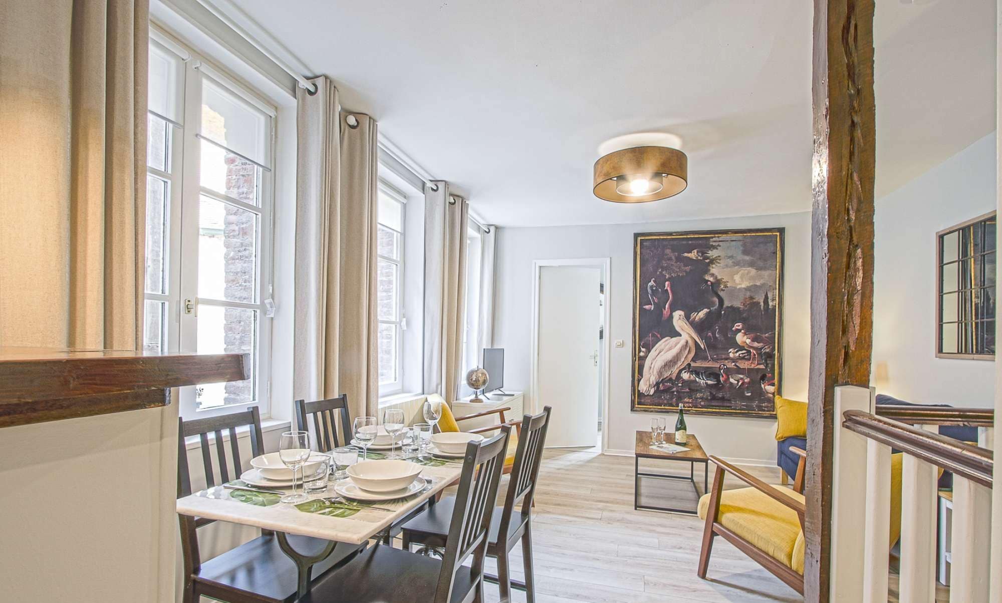 A dining area with a table set for four, a globe, and a bottle on the table. There is a large painting on the wall and a staircase to the right.