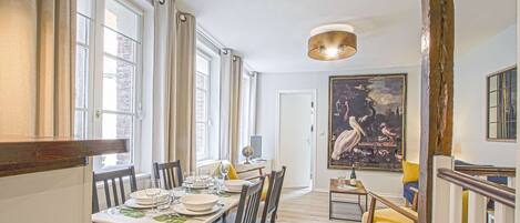 Un coin repas avec une table dressée pour quatre personnes, un globe terrestre et une bouteille posée sur la table. Un grand tableau orne le mur et un escalier se trouve à droite.
