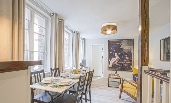 Un comedor con una mesa preparada para cuatro personas, un globo terráqueo y una botella sobre la mesa. En la pared hay un cuadro grande y una escalera a la derecha.