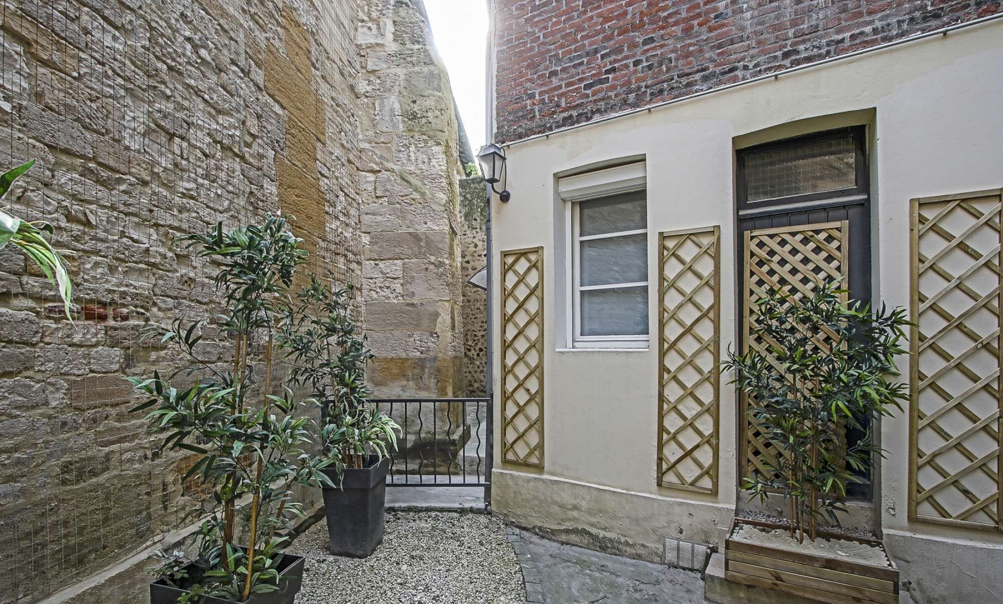 Een smalle steeg met een stenen muur aan de ene kant en een gebouw met een raam en een lattenpoort aan de andere kant. Op de voorgrond staan potplanten.