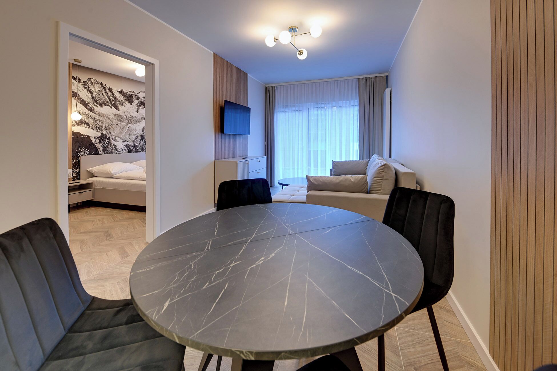 A modern, well-lit room with a round marble table, two black chairs, a sofa, and a TV mounted on the wall.