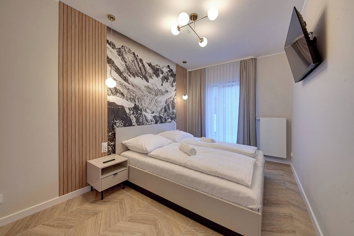 Une chambre à coucher moderne avec un grand lit, une table de chevet, un téléviseur fixé au mur et une fresque représentant un paysage de montagne.