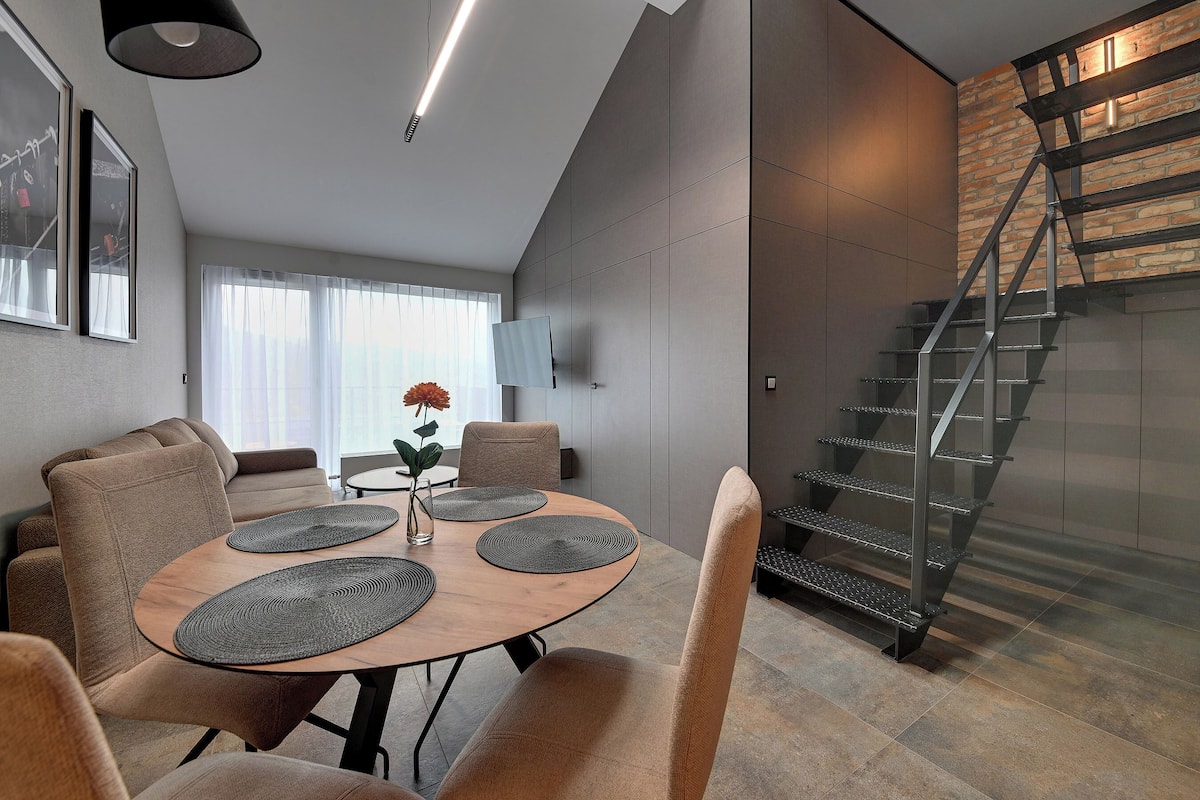 Un espace de salle à manger moderne avec une table ronde, des chaises rembourrées et un escalier doté d’une rampe en métal.