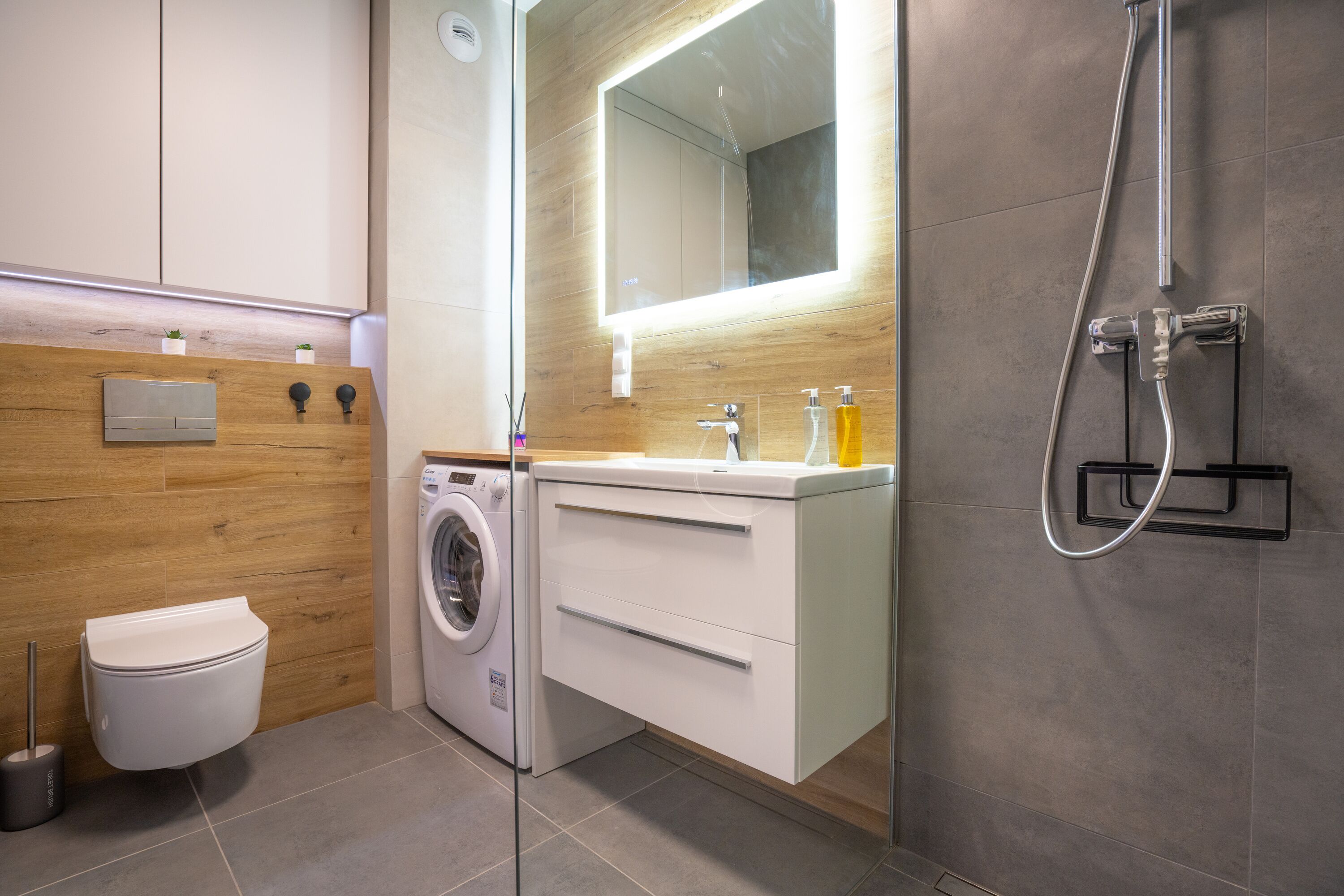 A modern bathroom with a built-in washing machine, a toilet, a sink with a mirror, and a shower.