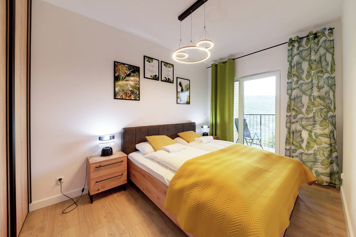 Une chambre à coucher comprenant un lit, des tables de chevet, une commode, un luminaire suspendu et une vue sur l’extérieur par une fenêtre ornée de rideaux.